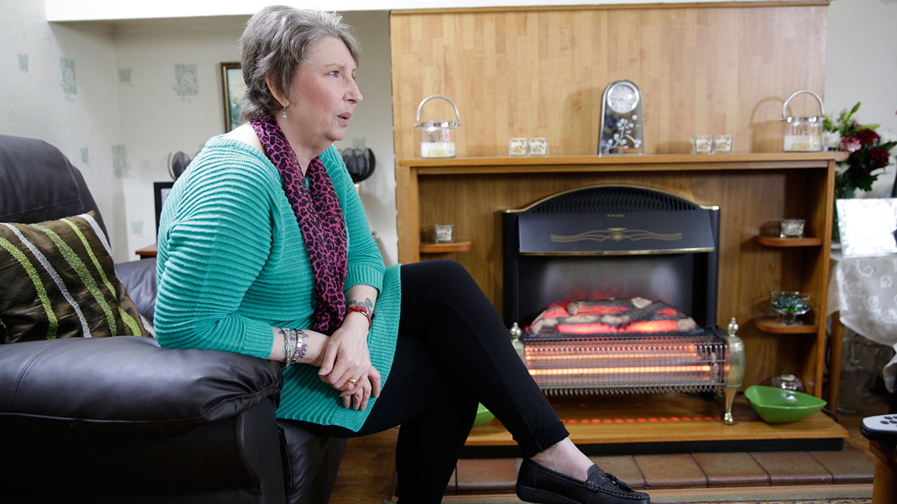 Cath is seated in a chair legs crossed. She is in her living room at home.