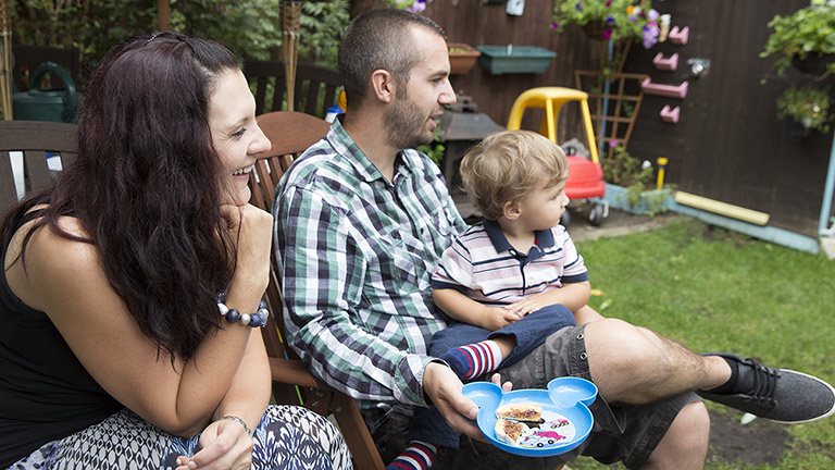 Luke and Lisa sit in their garden. They watch their little girl playing and their son sits on Luke's knee.