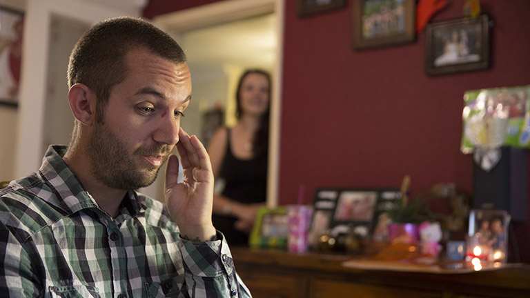 Luke sits in his living room. His wife Lisa watches him from the doorway in the background.