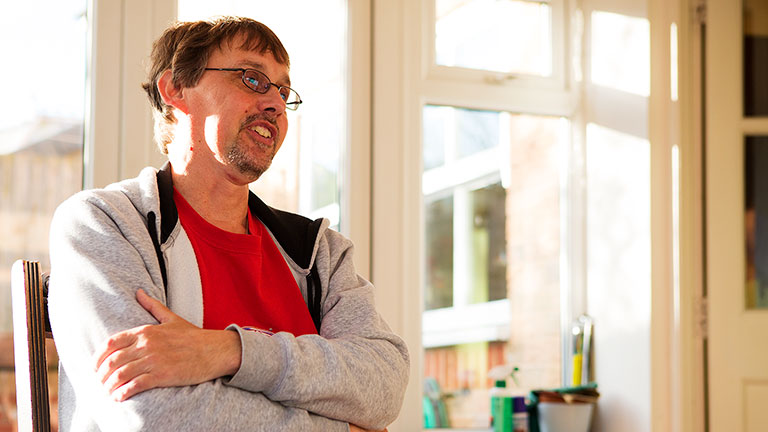 Paul stands with his arms crossed by a window in his home