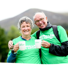 Man and woman in green Macmillan t shirts, holding Macmillan coffee mugs outside with a large hill in the background