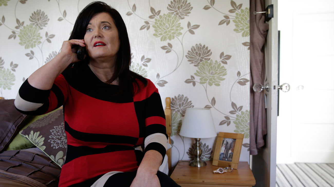Cathy sitting on a bed holding a phone to her ear.