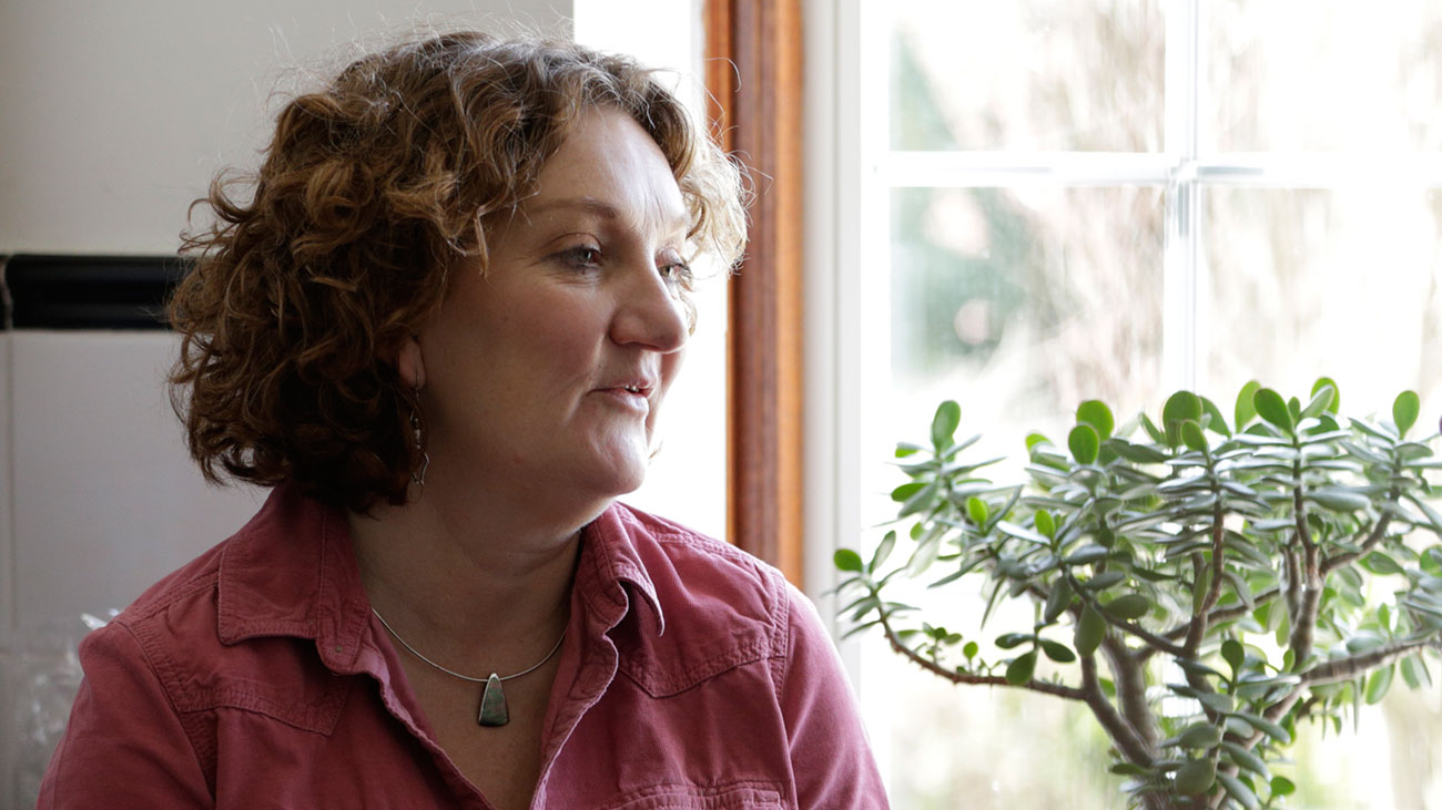 Daloni sits side-on in front of a window in an interior setting. There is a house plant behind her.