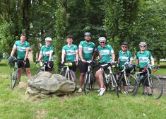 7 cyclists standing wearing green Macmillan cycling jerseys, standing next to their bikes on the side of the road.