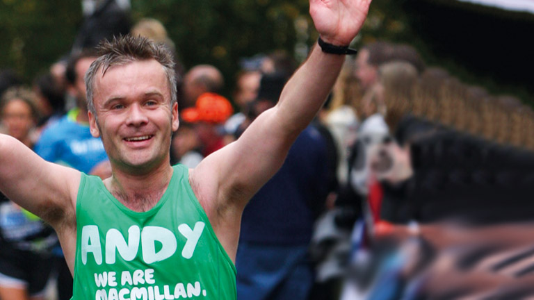 A man in a Macmillan running vest with Andy written on the front waves to the crowd during a race