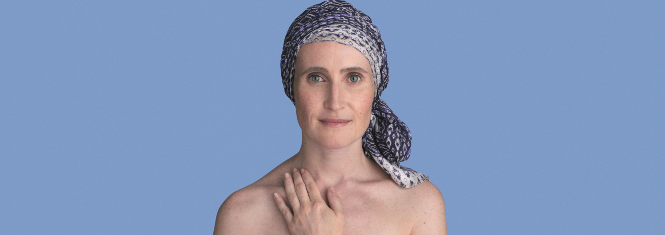 A close photograph of a woman wearing a headscarf is seen against a blue backdrop. She is not wearing a top and is resting one hand on her chest while looking directly at the camera.