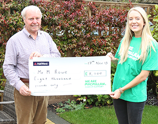 An older man wearing shirt and trousers and young woman wearing a green Macmillan vest holding up a giant cheque.