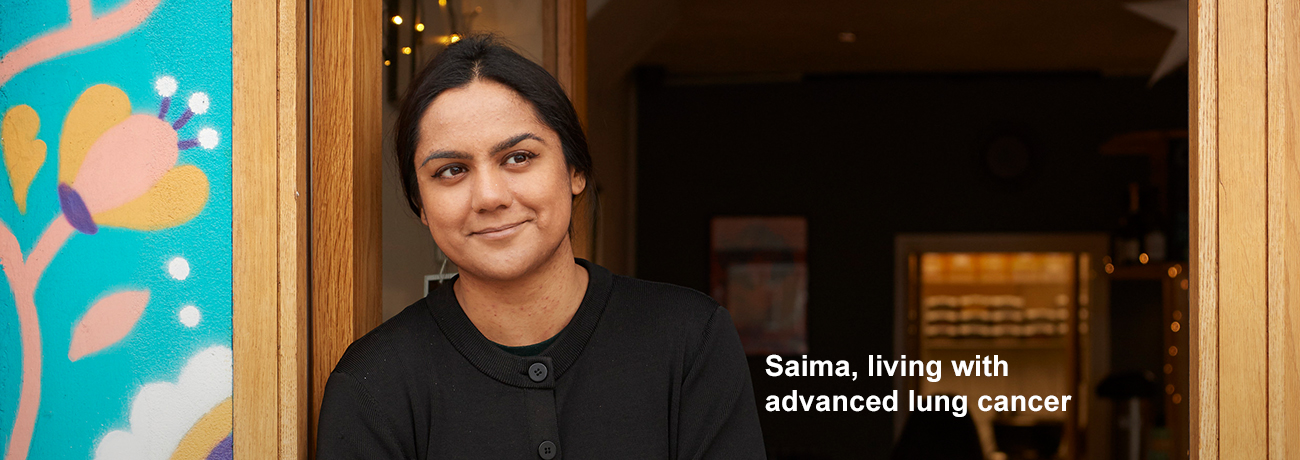 Saima, living with advanced lung cancer, smiles while leaning against a shop door. The caption next to her says, 'Saima, living with advanced lung cancer.'