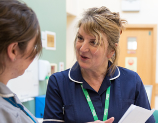 A female nurse wearing a green Macmillan lanyard is speaking to another woman.