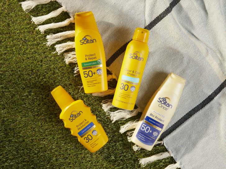 A photograph of Soltan products on the grass, in the sun. There is a black and white blanket on the grass where the yellow Soltan bottles are sitting.
