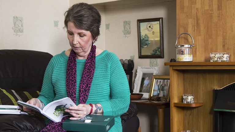 Cath reads a Macmillan booklet. She is seated in a chair in her home.