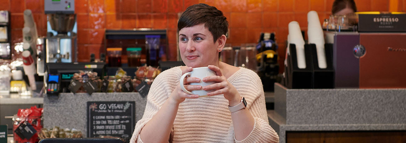 A woman with short cropped dark hair sitting in a café. She is holding a white mug with both hands.