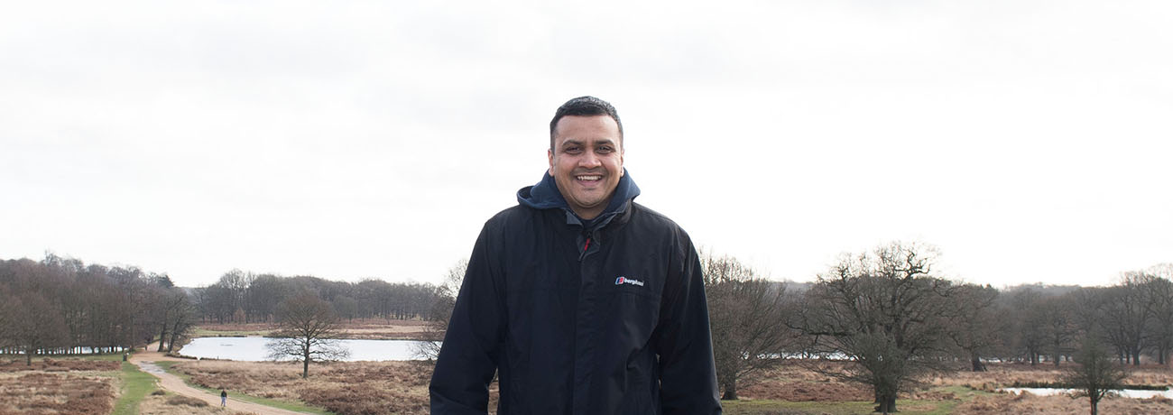 A man with short black hair wearing a black zip up jacket. He is standing in front of grass, lakes and trees without leaves.