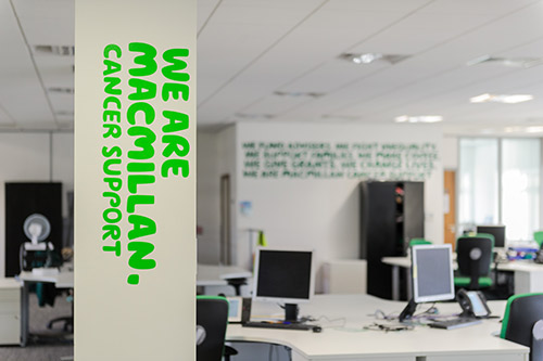 Internal view of Macmillan office with computers and desk in the background the the words 'We are Macmillan. Cancer support' on the pillar in the foreground.