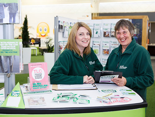 A Macmillan staff member sits at an information stand in Boots, there are lots of leaflets and cancer information booklets on the table.
