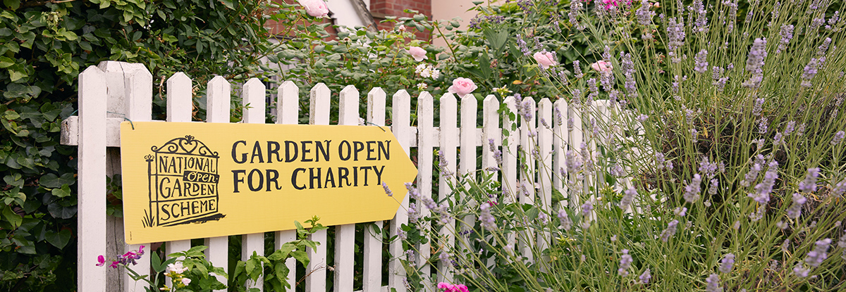 A yellow sign that says 'Gardens open for charity' in black letters. The sign is on a white fence surrounded by flowers.