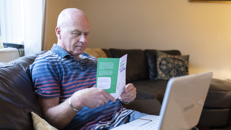 A man sits on a sofa reading a cancer information leaflet, a laptop is open on his lap.