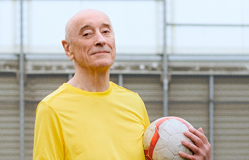A man poses for a picture on a football pitch. He is holding a football and wearing a yellow t-shirt.
