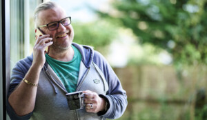 Stewart is leaning against a door frame. He is smiling, holding a mobile up to his ear and has a mug in his other hand. 