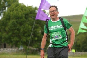 Richard is standing outside and wearing a green Macmillan top. He has on a rucksack and pinned to his top is his registration number.