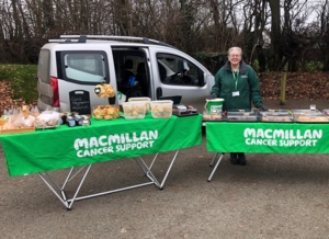 Irene is outside and standing behind a table that is lined up with baked goods. She is wearing a green Macmillan jacket and has her hand placed on a Macmillan bucket.