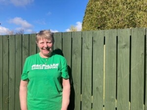 Olywyn is facing the camera and leaning against a faded green face. She is wearing a green Macmillan tshirt and smiling.