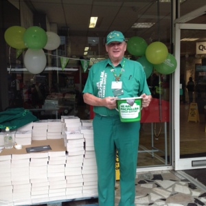 Brian is standing in a full Macmillan green outfit and is holding a Macmillan bucket. He appears outside and is standing in front of a large pile of books. 