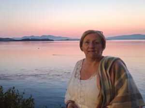 Shirely is outside and behind her is a body of water. She is looking at the camera and is wearing a white top. Draped over her right shoulder is a shall. 