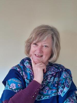 Liz is smiling at the camera and has her hand with a finger extended rest on her chin. She is wearing a blue patterned shall around her shoulders.