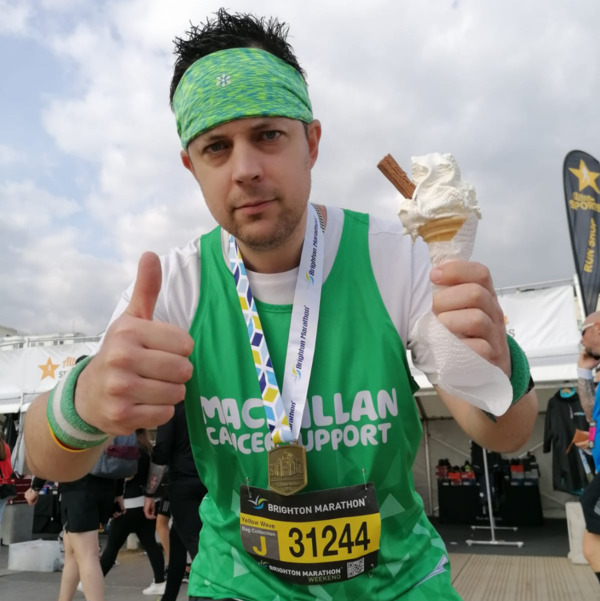 Robbie is standing and leaning towards the camera. In his right hand he is holding an ice cream and in his left is giving a thumbs up. He is wearing a Macmillan running top.