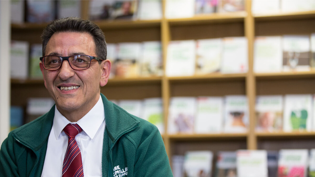 Mario, a middle-aged man wearing glasses, is sitting in front of shelves with cancer information booklets.