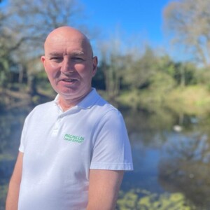 Calum is looking at the camera and appears to be outside. He is wearing a white polo top that has a Macmillan logo on it. 