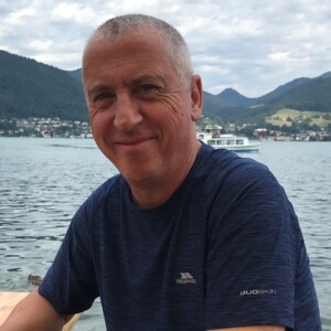 Joe appears outside near a body of water. He is looking at the camera and smiling. He is wearing a dark blue top. 