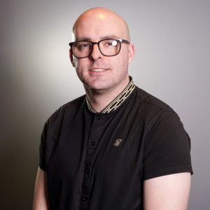 Daniel is looking at the camera. He is wearing a black top and black framed glasses.