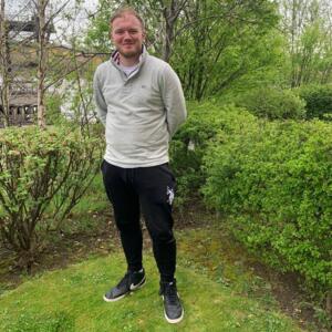 James is outside in what appears to be a garden. He is smiling and looking at the camera. He is wearing black trousers and a white cream jumper. 
