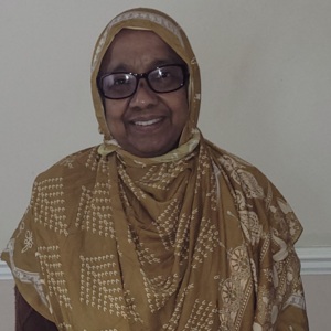 Sarifa is standing and smiling at the camera. She is wearing a brown and white patterned head scarf and black trimmed glasses.