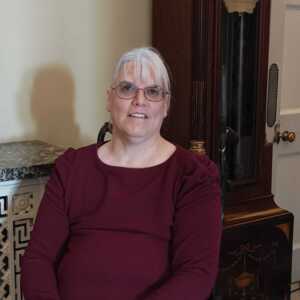 Fiona is sat down and looking at the camera. She is wearing a maroon top and glasses. 