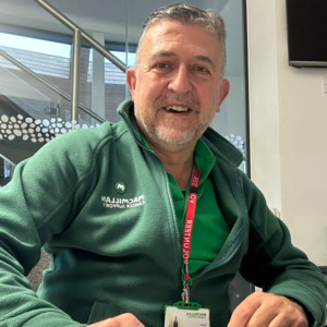 A selfie of Victor where he is sat a desk looking at the camera. He is wearing a green Macmillan jumper and appears to be in front of a computer.