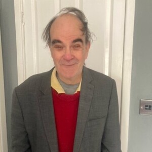 Duncan is standing and looking at the camera. He is wearing a grey jacket and a red jumper.