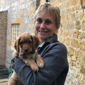 Joe is seen outside and is smiling at the camera. She is wearing a blue grey jumper and is holding a puppy in her hands. 