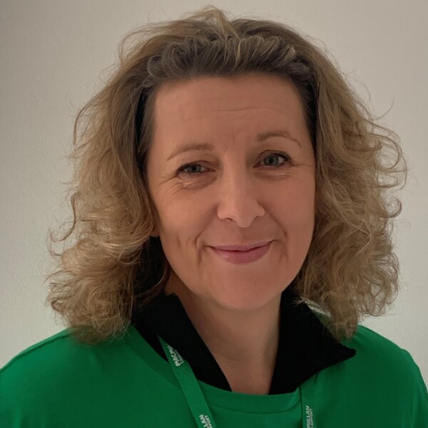 Claire is looking at the camera and smiling. She is wearing a green Macmillan t-shirt and lanyard.