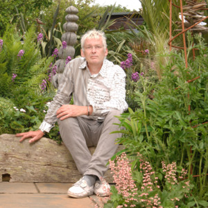 Geoffrey is outside and sat in a garden. He is looking at the camera and is wearing a white patterned shirt and tan trousers.