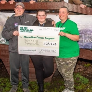 Siobhan is seen standing and holding a large Macmillan cheque. To her left is a man and to her left is a woman wearing a green Macmillan top. They are standing outside.