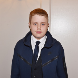 Jake is standing and looking at the camera. They are wearing a dark blue jacket, dark blue tie and a white shirt.