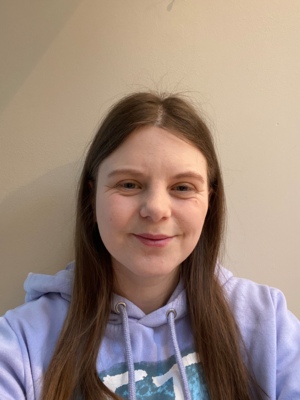 A selfie of Sarah from the shoulders up. She is looking at the camera and smiling. She has long brown hair and is wearing a bright purple jumper.