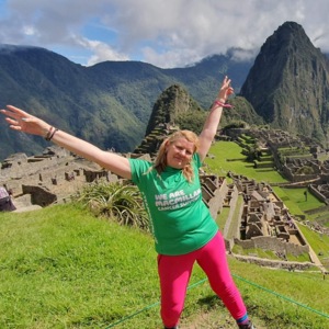 Clare is standing outside with her arms extended. Behind her is Machu Picchu. She is wearing a green Macmillan top and bright pink trousers.