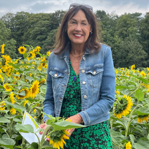 Judy is standing in a field of sunflowers. She is smiling and looking at the camera. She is wearing a denim jacket and a dress with a green flower pattern. 