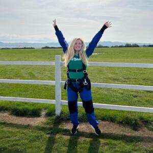 Tasmin is standing outside with her arms in the air. She is smiling at the camera and appears to be wearing a skydiving outfit. 