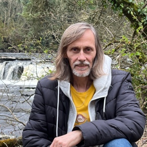 Paul is outside and is sat down near a waterfall. He is looking at the camera and wearing a yellow top, gray hoodie and black jacket.