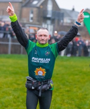 Gary is outside and holding his arms in the air. He is wearing a Macmillan top and appears to be in running gear.